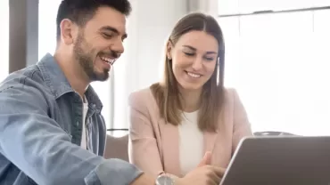 Two people are sitting at a table facing a laptop while smiling.