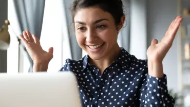 Person smiling and looking at a laptop, raising hands in excitement.