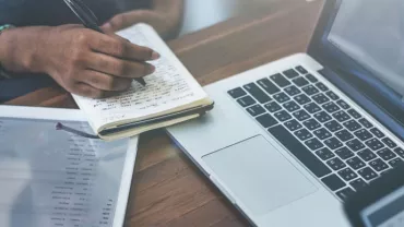 Person writing in notebook next to a laptop and tablet.