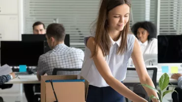 Person arranging desk with plant in a busy office setting.